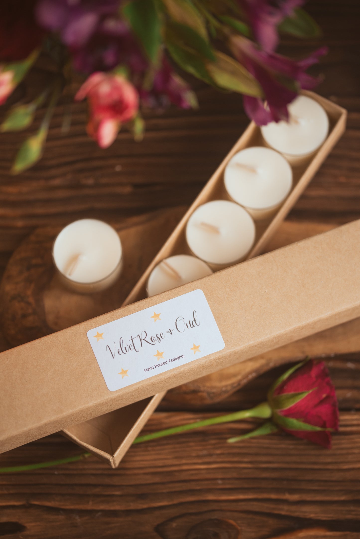 Set of white candles in a cardboard holder with a label on a wooden surface, surrounded by flowers and a rose.