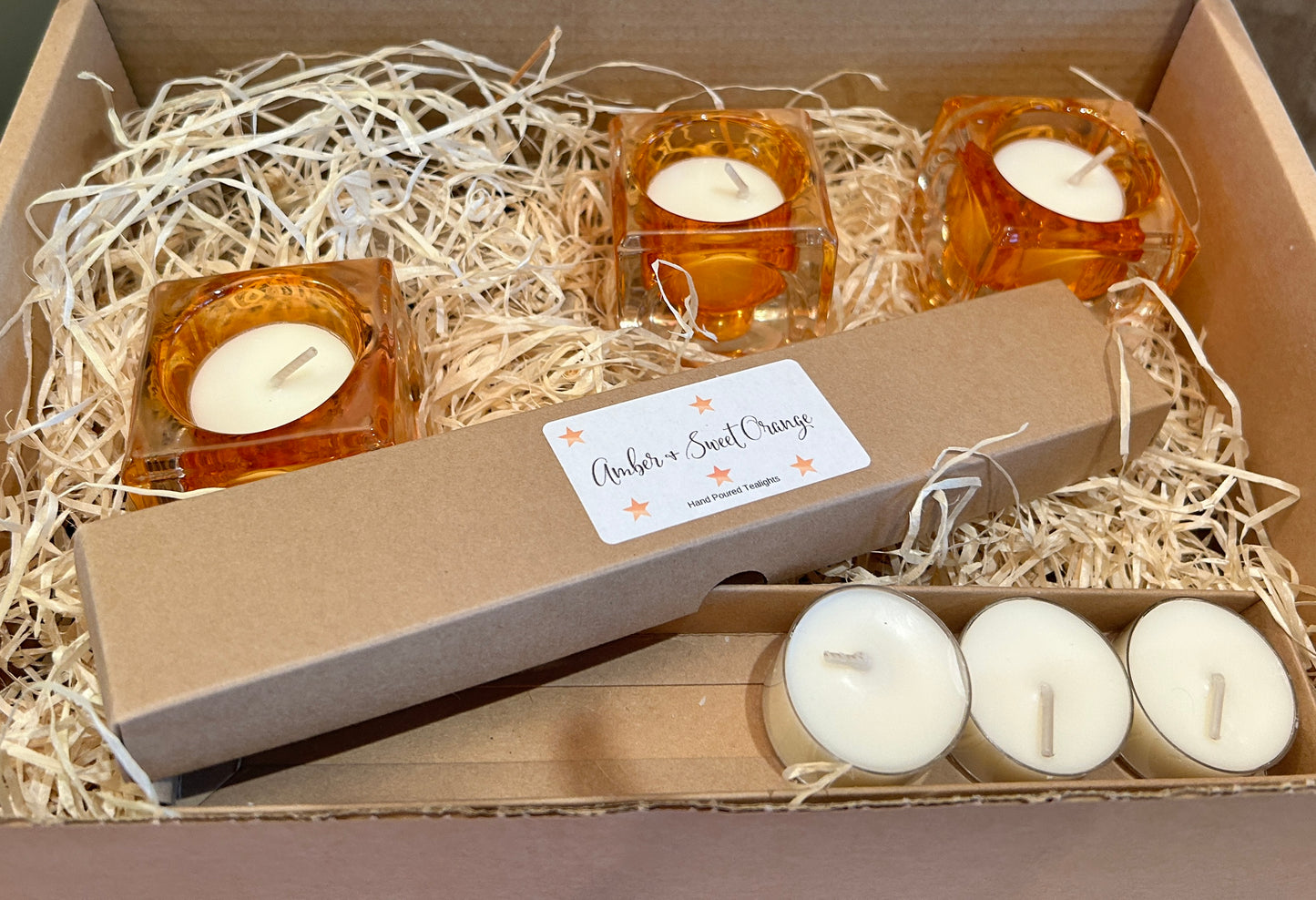 3 x orange square glass tealight holders with tealight candles in a box with wooden shredded packaging.