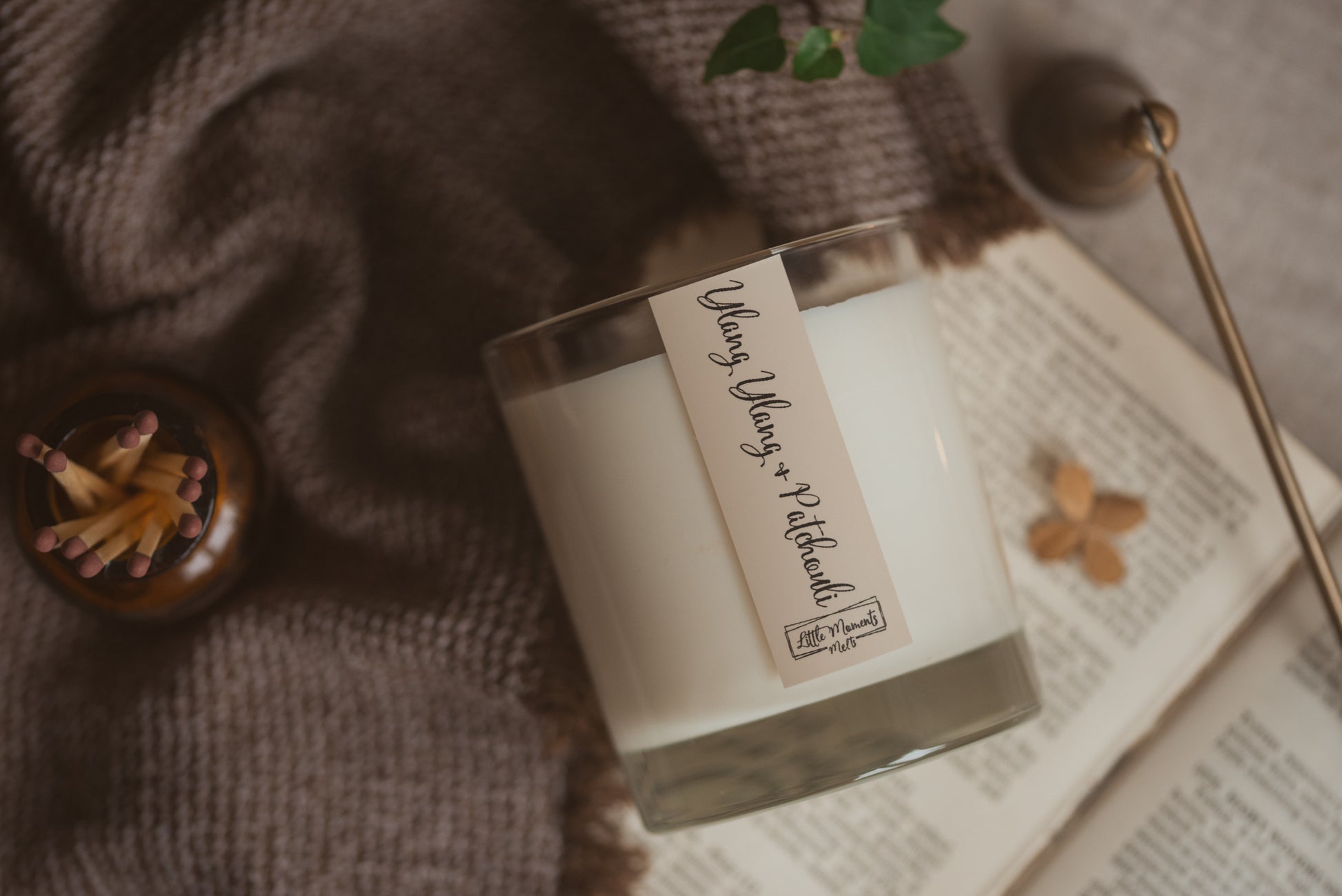 candle on its side with. label saying ylang-ylang and patchouli, surrounded by matches, hessian sheet and a book 