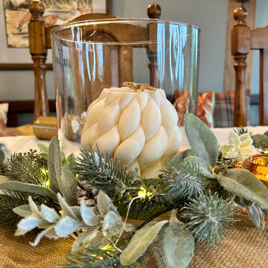 large braided white candle in glass  jar with garland around it 