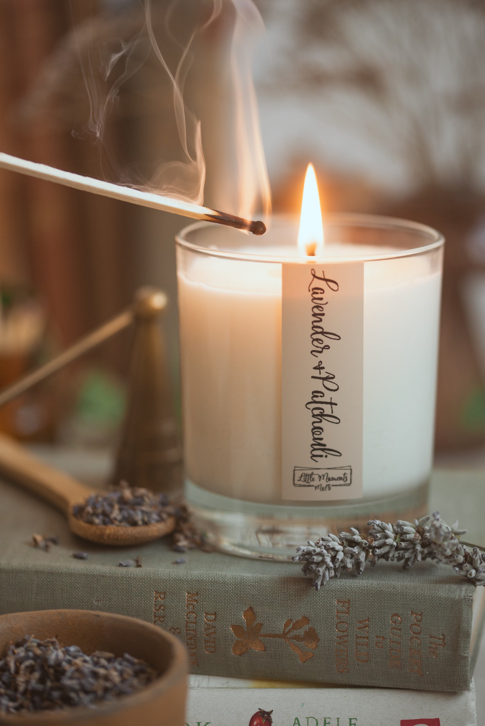 lit candle with lit match near the wick, the label reads lavender and  with lavender flowers around the base. 