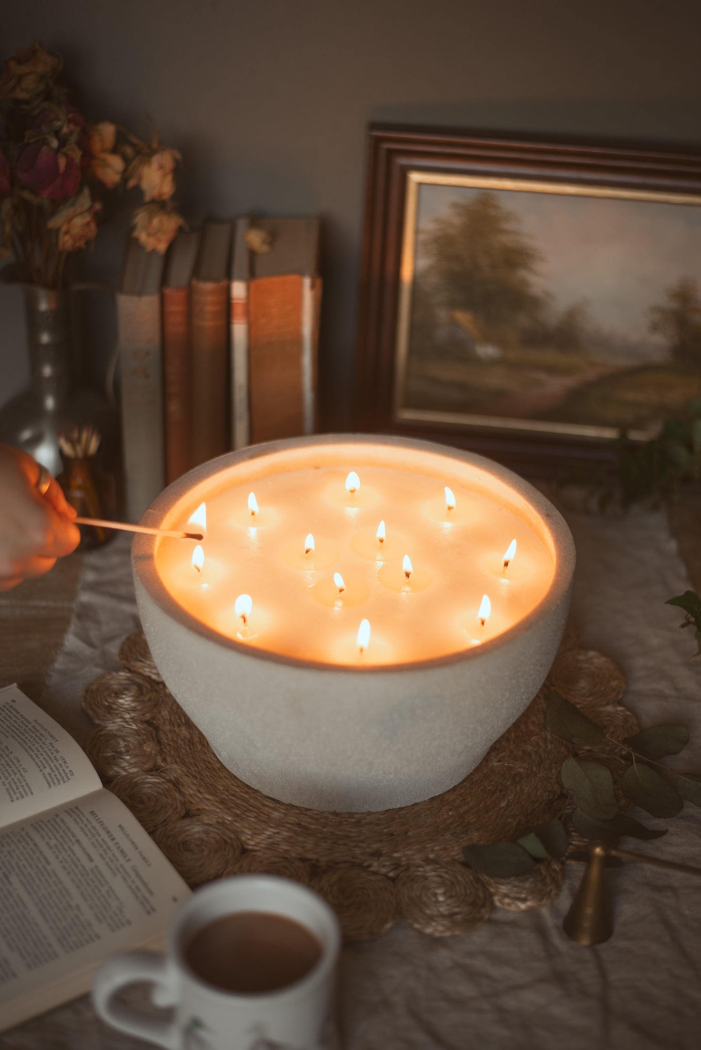 large bowl candle with 12 wicks all lit, beside is a mug, picture frame, old books and flowers in a country cottage style 