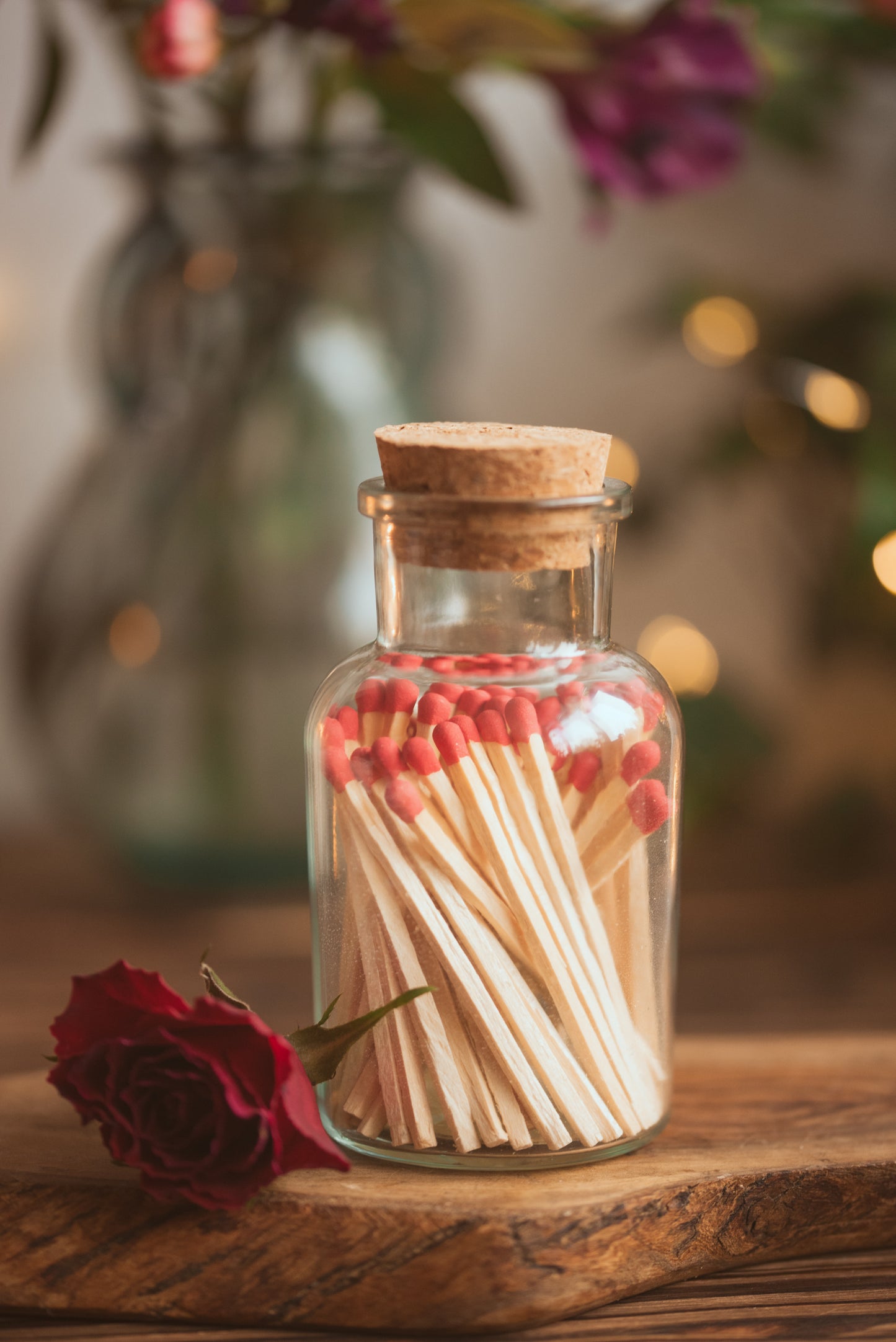 Jar of matches with a cork lid on a wooden surface, surrounded by flowers and lights.