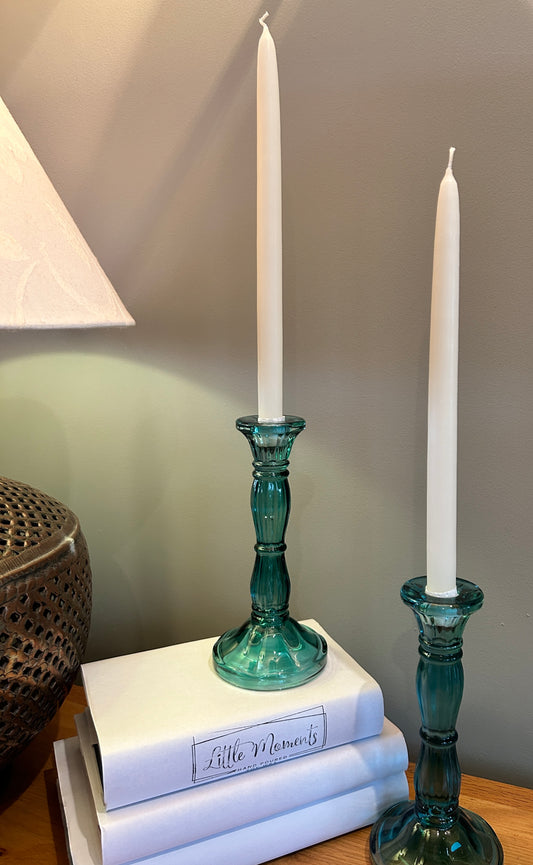 Two tall blue glass candlesticks with tapered dinner candles, beside books and lamp 