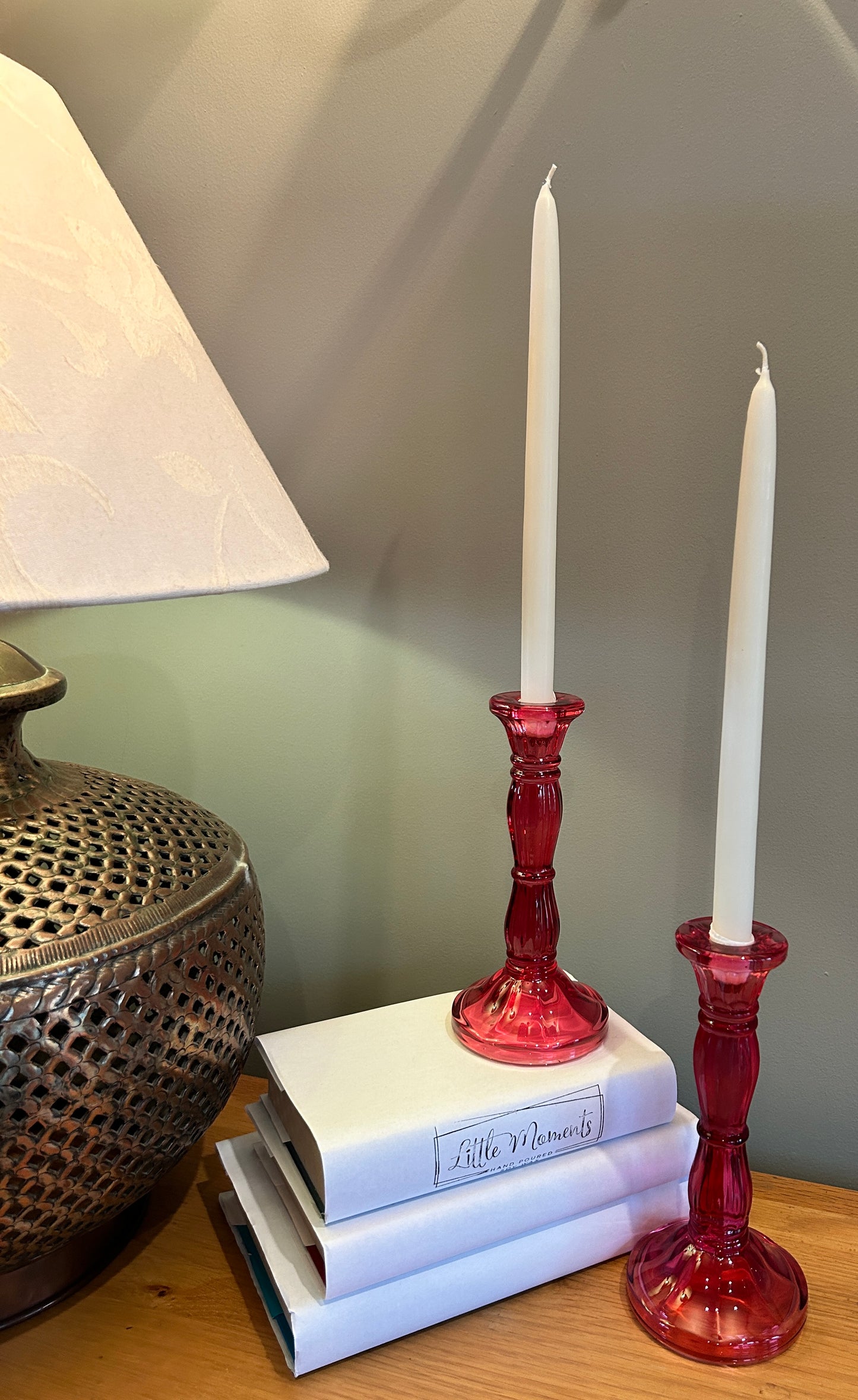 Two pink glass candlesticks with tapered dinner candles, beside a lamp and books 