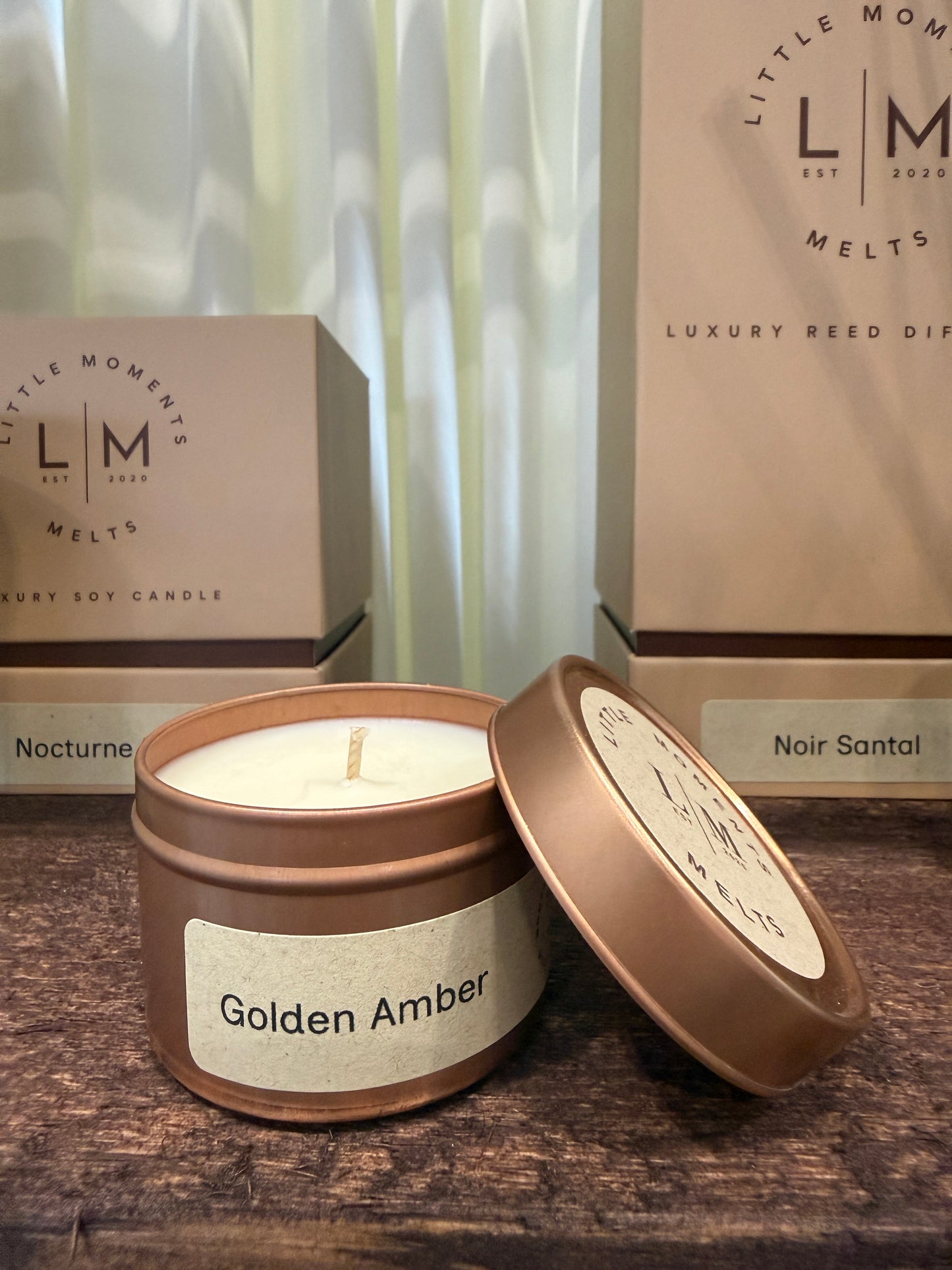 Two candle tins labeled 'Golden Amber' and 'Nocturne' with 'LM Moments Melts' branding on a wooden surface.
