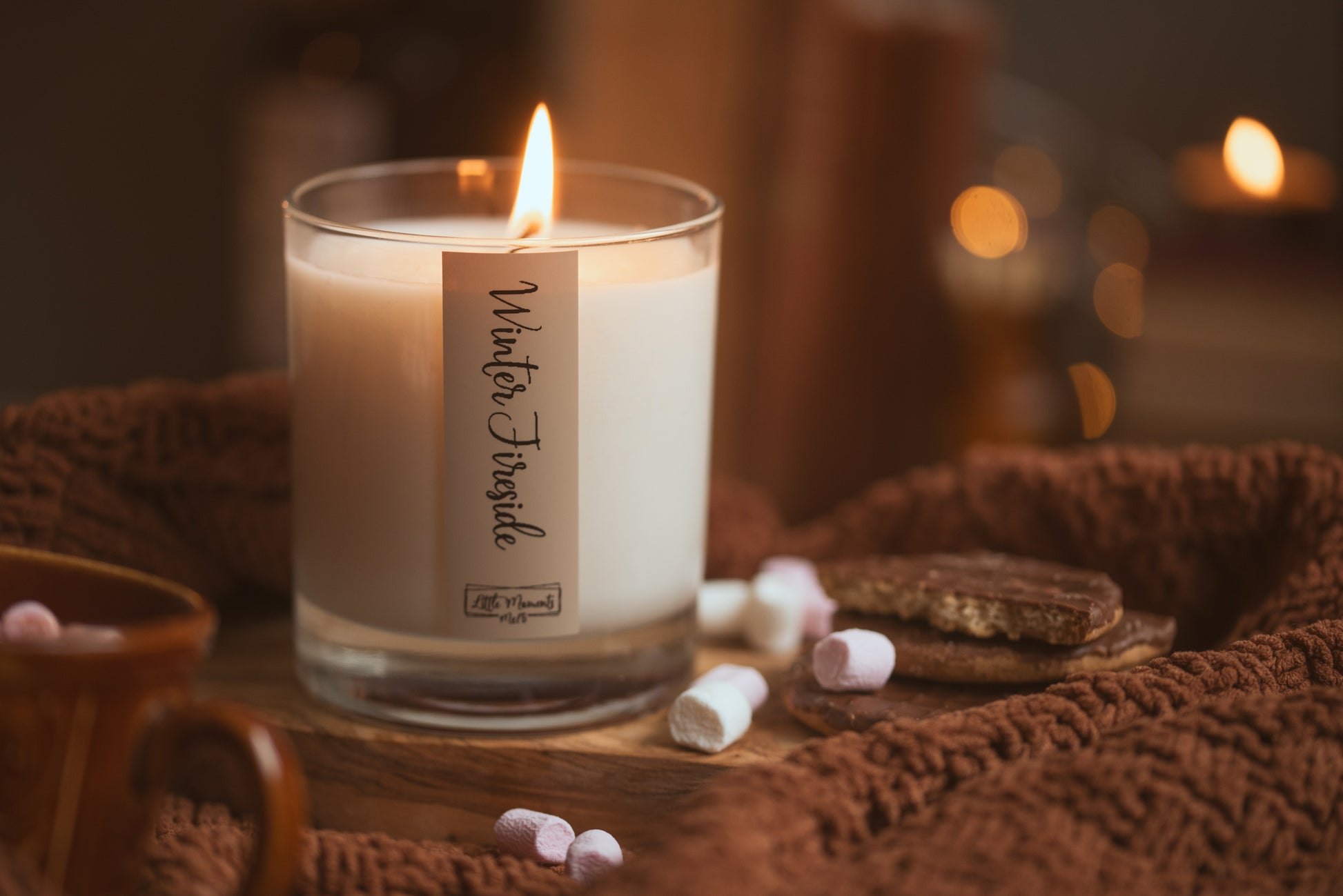 Candle labeled 'Winter Twinkle' on a woven surface with marshmallows and cookies.